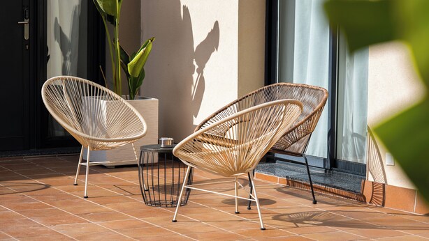 Twee rieten stoelen op een betegeld terras met een plant in pot en een metalen tafel.