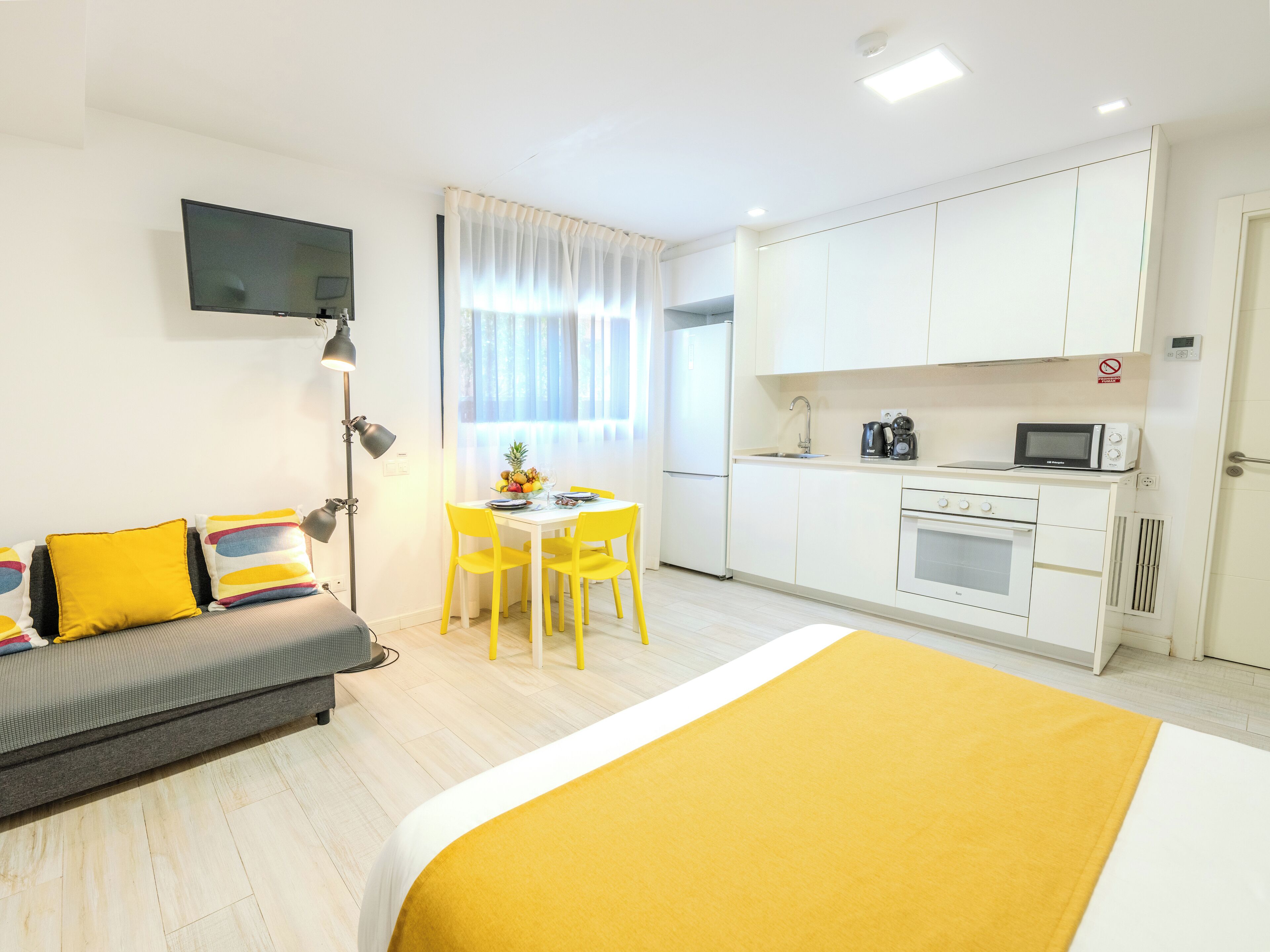 A modern hotel room with a bed, a sofa, a dining table with chairs, a kitchen area, and a flat-screen TV mounted on the wall.