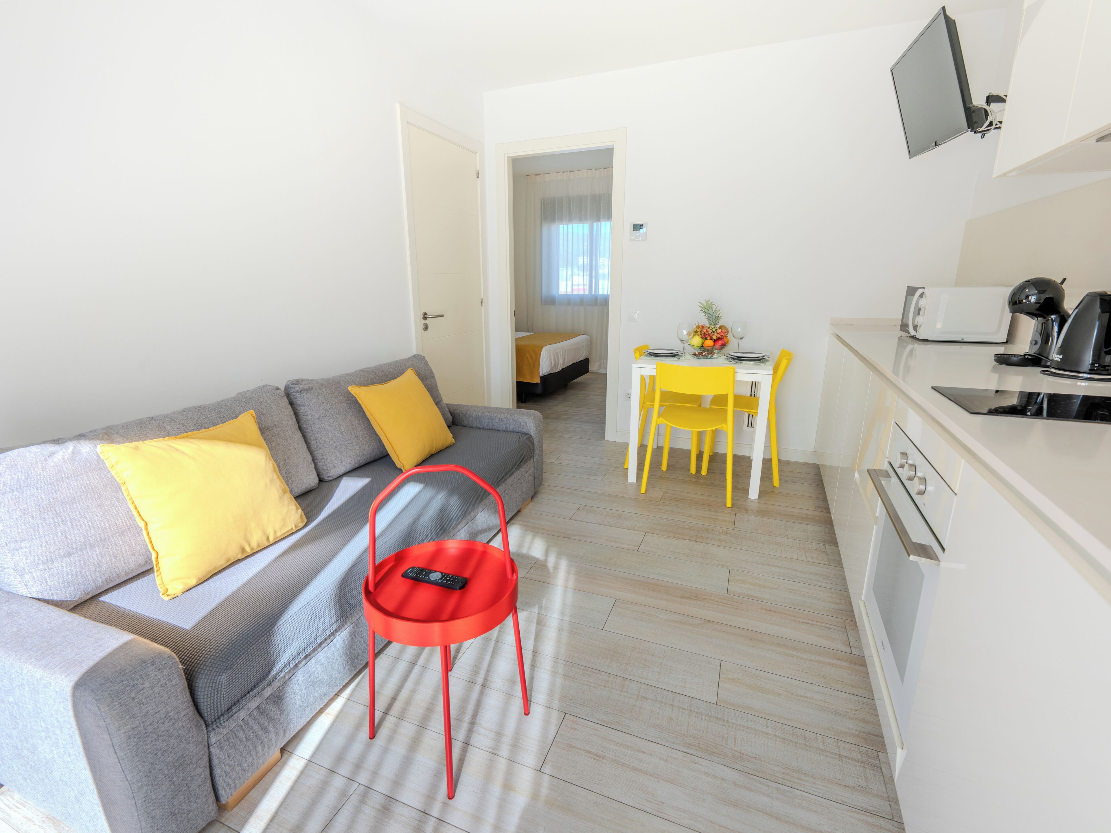 A modern living room with a grey sofa, yellow cushions, a red chair, a dining table with yellow chairs, and a kitchen area with a microwave and toaster.