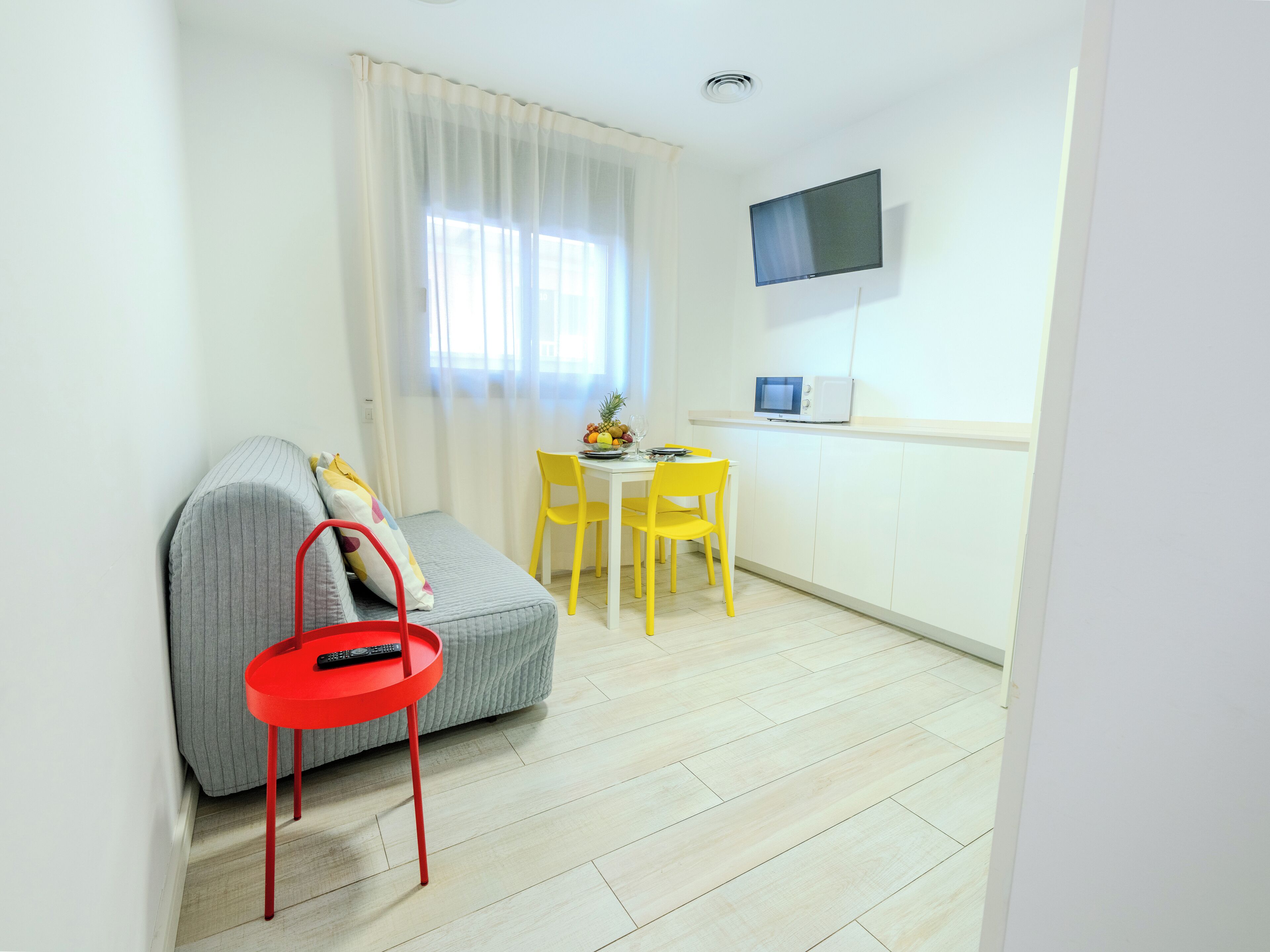 Un espacio habitable compacto con un sofá, una pequeña mesa de comedor con sillas amarillas, una mesita auxiliar roja y un televisor de pantalla plana montado en la pared.