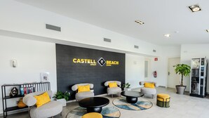Een hotel lobby met een receptiebalie, een zithoek en een groot bord waarop 'CASTELL BEACH HOTEL' staat.