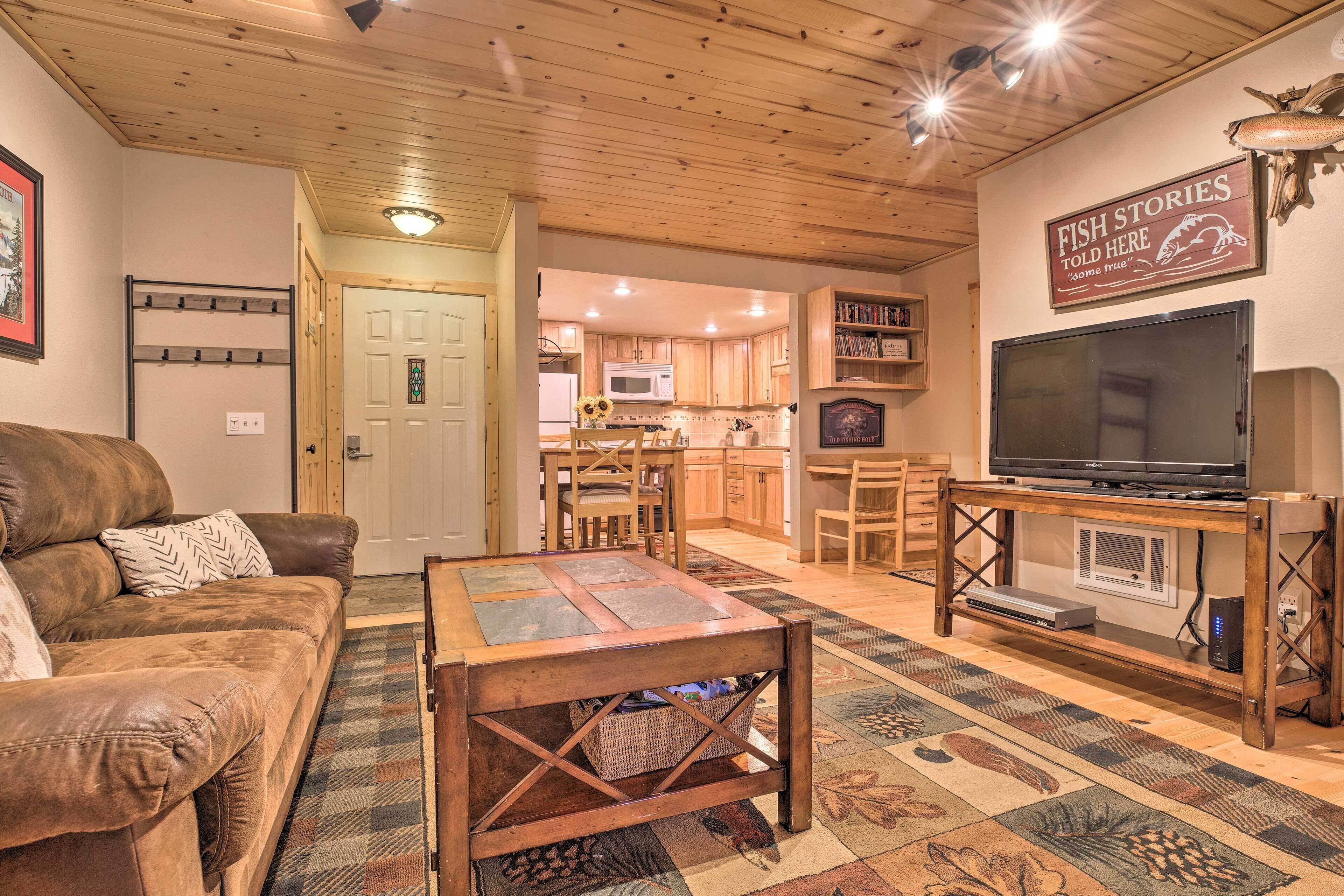 A cozy living room with a leather couch, a coffee table, and a TV on a wooden stand. There is a kitchen in the background with wooden cabinets and a microwave.