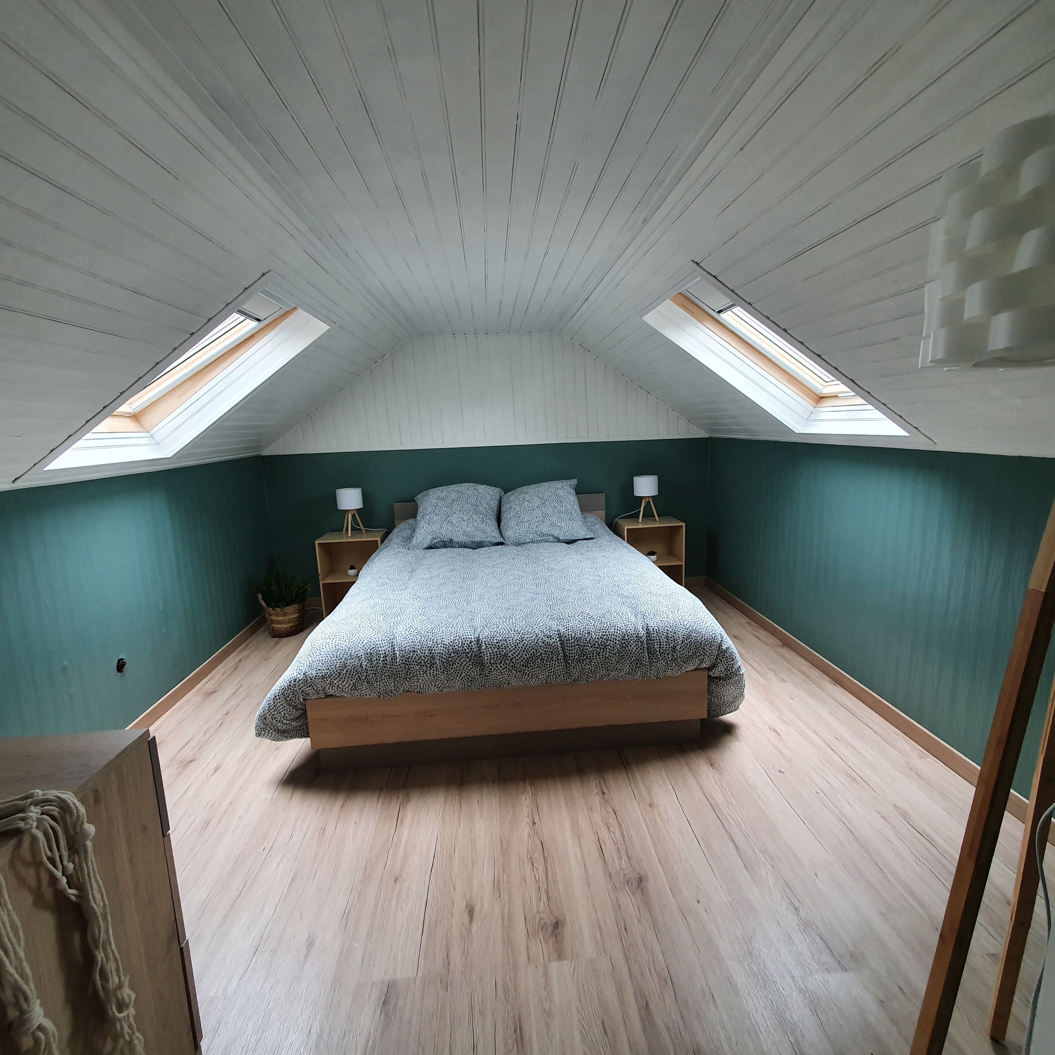Ein Schlafzimmer mit schrägen Wänden, Dachfenstern, einem Bett mit gemusterten Betttextilien, Nachttischen mit Lampen und einem Holzboden.