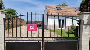 Une entrée fermée par un portail, agrémentée d’une pancarte de bienvenue, une maison avec un toit en tuiles et une allée gravillonnée.