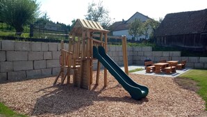 Un parque infantil con un tobogán de madera y estructura para escalar, rodeado por un muro de piedra y una zona de mesas de picnic.