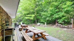 Eine Holzterrasse mit Picknicktischen und Grillplatz, umgeben von einem bewaldeten Garten.