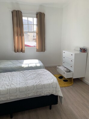 A bedroom with a bed, a dresser, a window with curtains, and a view of buildings outside.