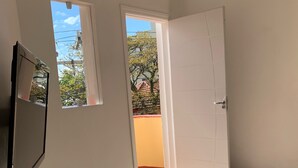 Habitación con piso de madera, un escritorio blanco con control remoto, una televisión montada en la pared y una puerta que da a un área exterior con vista a árboles y un edificio.