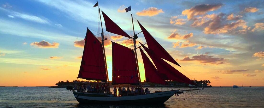 Een zeilboot met meerdere zeilen, een dek met mensen en een zonsondergang op de achtergrond.
