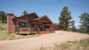 A two-story log cabin with a wooden deck and a chimney, surrounded by a gravel driveway and trees. - Perfect Mountain Home II with HOT TUB overlooking Mary's Lake/Near RMNP entrance (Estes Park)