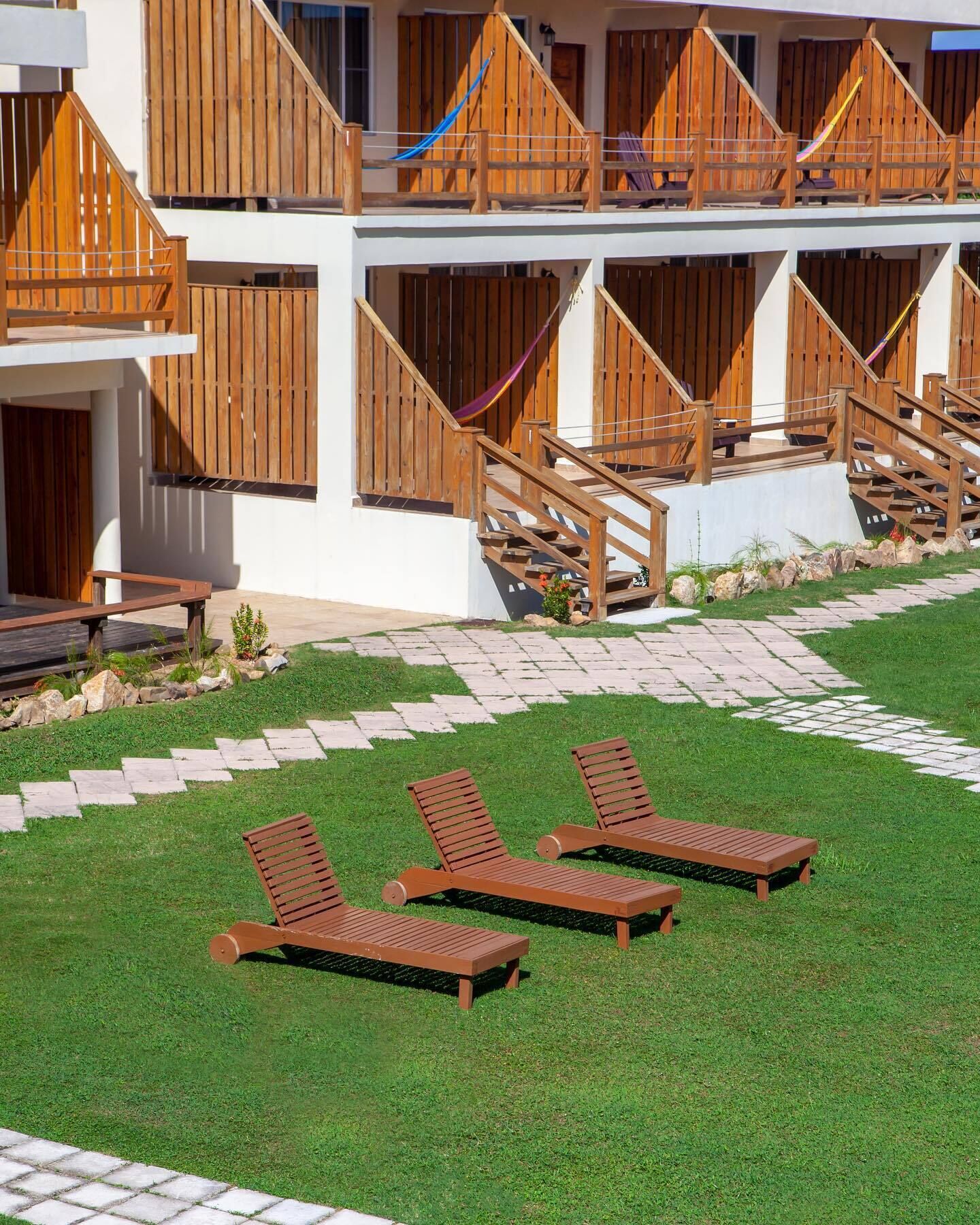 Un hotel con balcones de madera, un área de piscina con sillones de descanso y un camino de piedra.