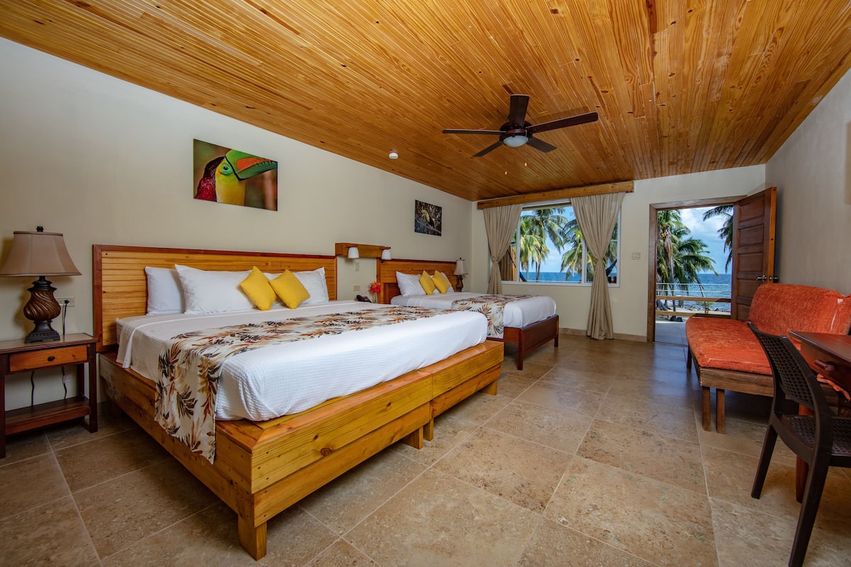 A room with two wooden beds, a wooden ceiling fan, a wooden table, and a view of the ocean.
