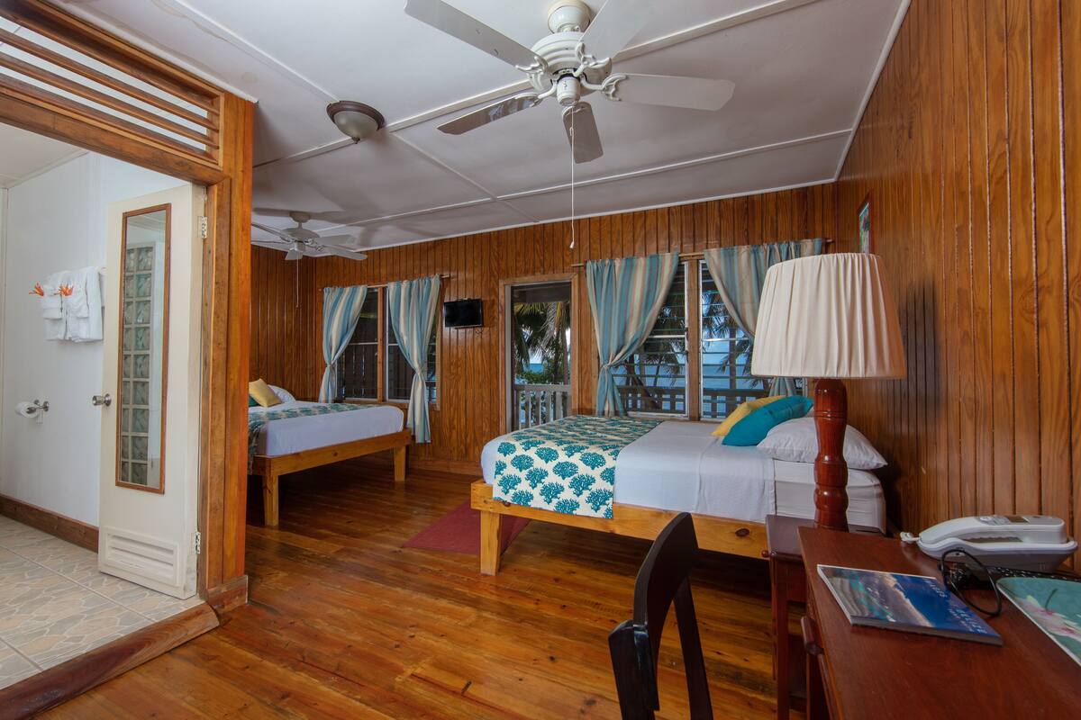 A room with two beds, a ceiling fan, wooden walls, a window with curtains, and a desk with a lamp.