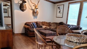 Una sala de estar acogedora con piso de madera, una mesa de comedor con cubierta de vidrio y un sofá. Se observan dos ciervos montados en la pared y un ventilador de techo.