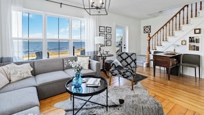 Un salon spacieux avec une grande fenêtre donnant sur la mer, un lustre moderne, un escalier en bois et un coin salon confortable.