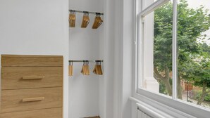 Une pièce aux murs blancs, avec une armoire en bois et une fenêtre donnant sur des arbres et un bâtiment en briques.