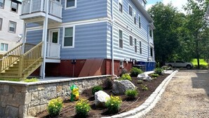 A two-story residential building with a balcony, a stone foundation, and a landscaped garden with rocks and plants.