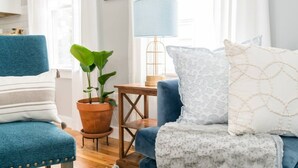 A living room with a blue armchair, a sofa with patterned cushions, a potted plant, a small wooden table, and a lamp.