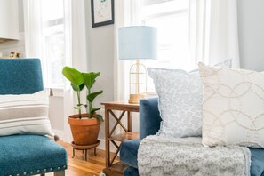 A living room with a blue armchair, a sofa with patterned cushions, a potted plant, a small wooden table, and a lamp.