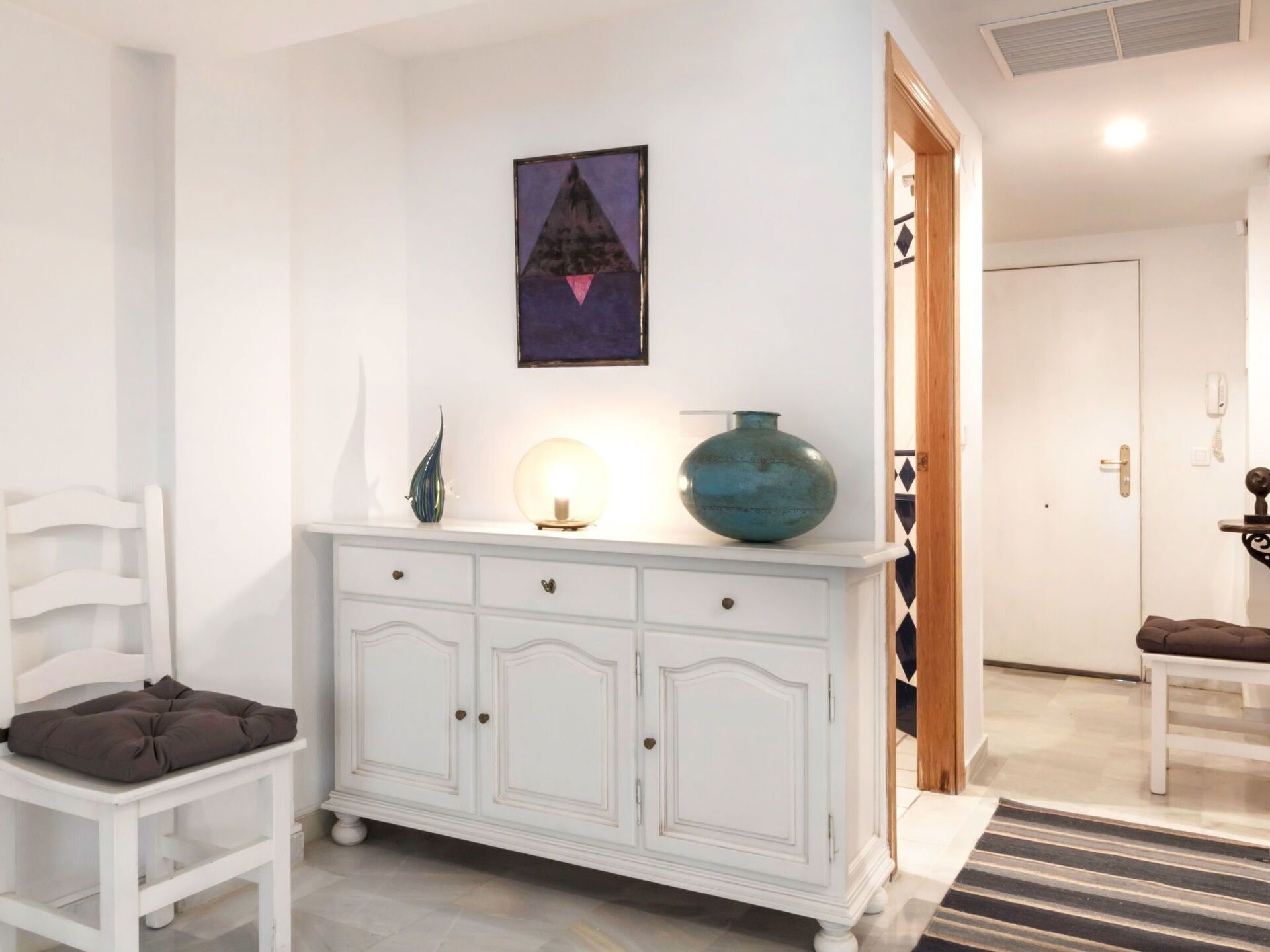 A white cabinet with drawers, a blue vase, a lit candle, and a painting on the wall.