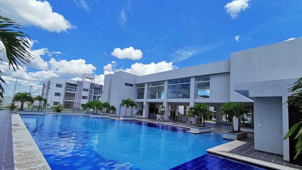 A modern hotel with a large swimming pool, multiple balconies, and a clear blue sky.