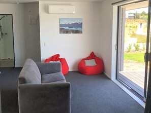 Une pièce moderne avec un canapé gris, deux fauteuils en forme de poire rouge, un tableau représentant des montagnes et une porte-fenêtre coulissante donnant sur un jardin.