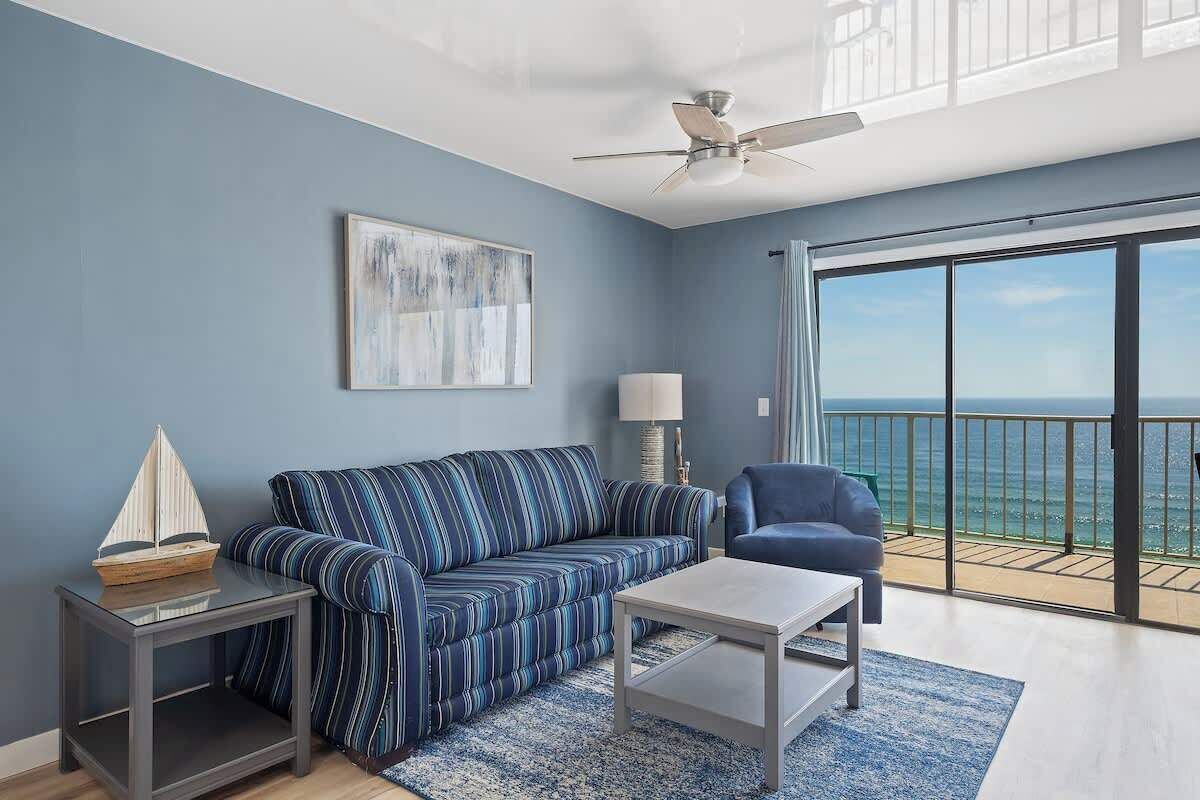A modern living room with a sofa, a coffee table, a side table, and a lamp. There is a balcony with a view of the ocean.