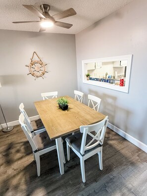 Un comedor con una mesa de madera, sillas blancas, un ventilador de techo y un espejo en la pared que refleja una cocina.