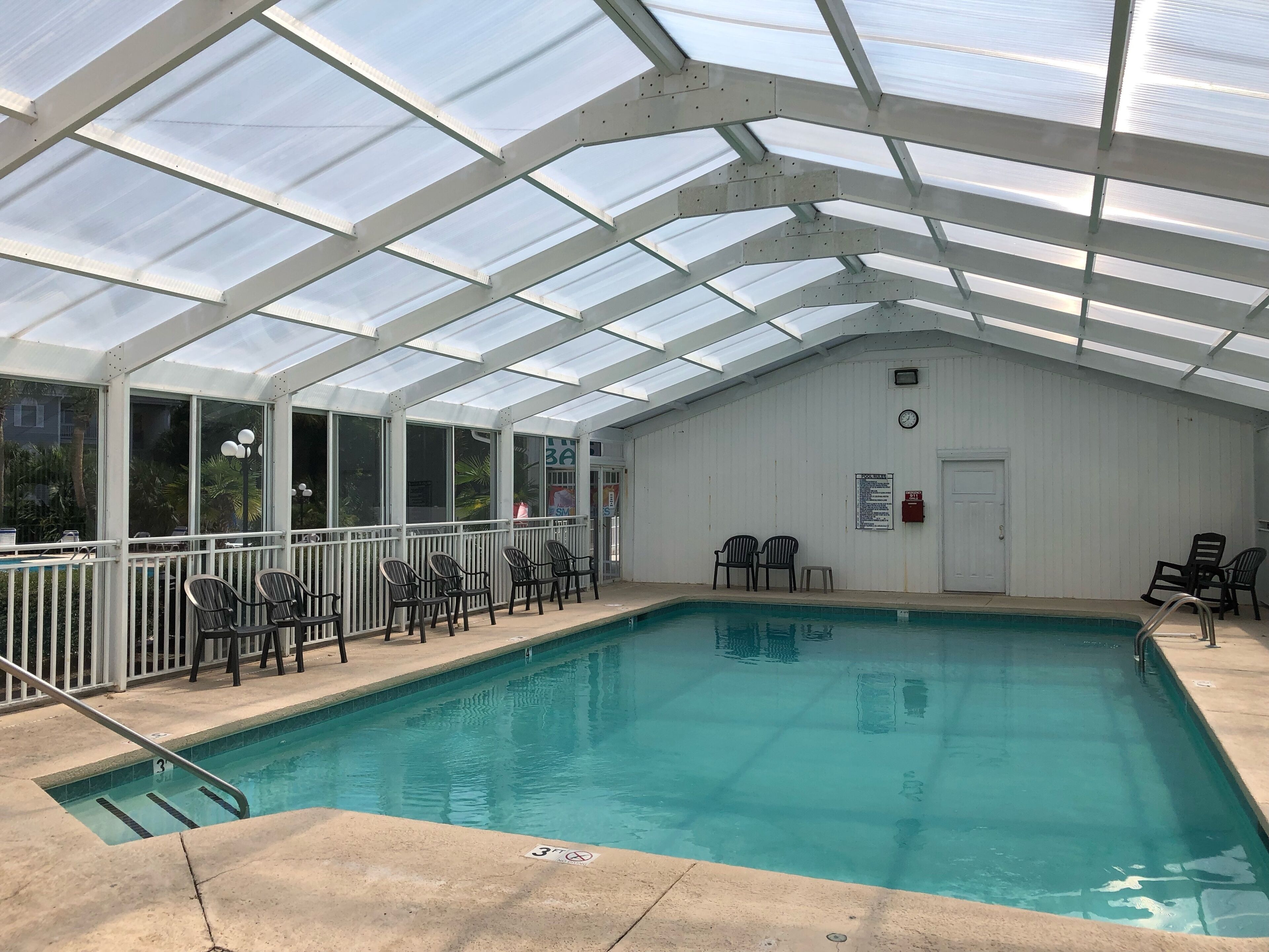 Piscina interna com teto de vidro, cercada por cadeiras e uma parede branca com um relógio.