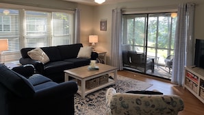 A living room with a black sofa, a white coffee table, and a patterned armchair. There is a ceiling fan, a lamp, and a view through large windows to the outdoors. - Sea Trail 2BR - Golf, Bike, Beach Getaway! Clean, Quiet, Pools, Great View! (Sunset Beach)