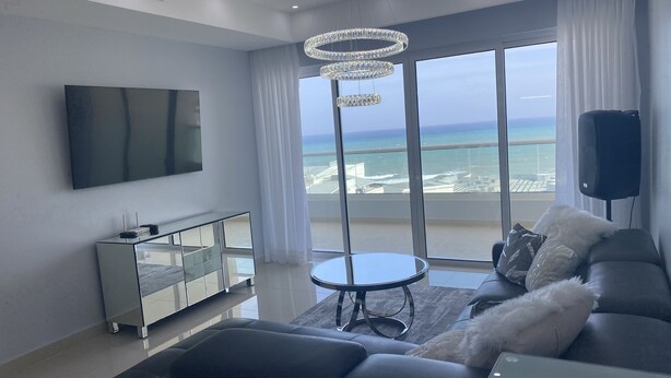 Ein modernes Wohnzimmer mit großem Fernseher, einer Glasschrank, einem runden Tisch, einer Couch und einem Balkon mit Meerblick.