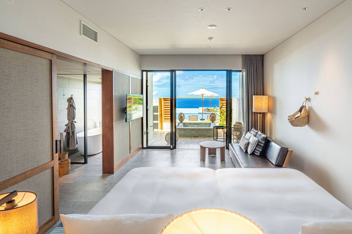A modern hotel room with a large bed, a balcony with a view of the sea, a seating area with a table and chairs, and a television.