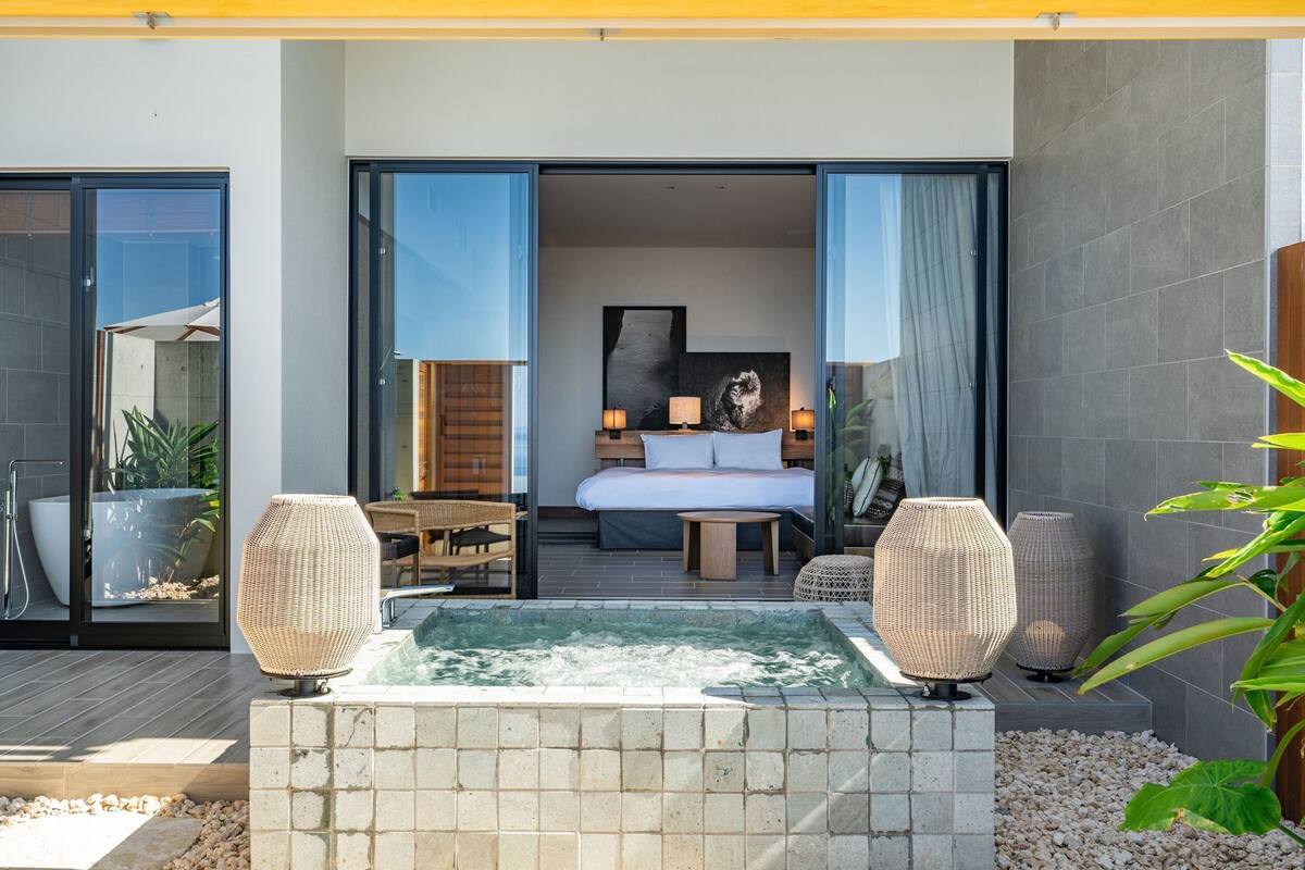 A modern hotel room with a large bed, a small seating area, and a hot tub.