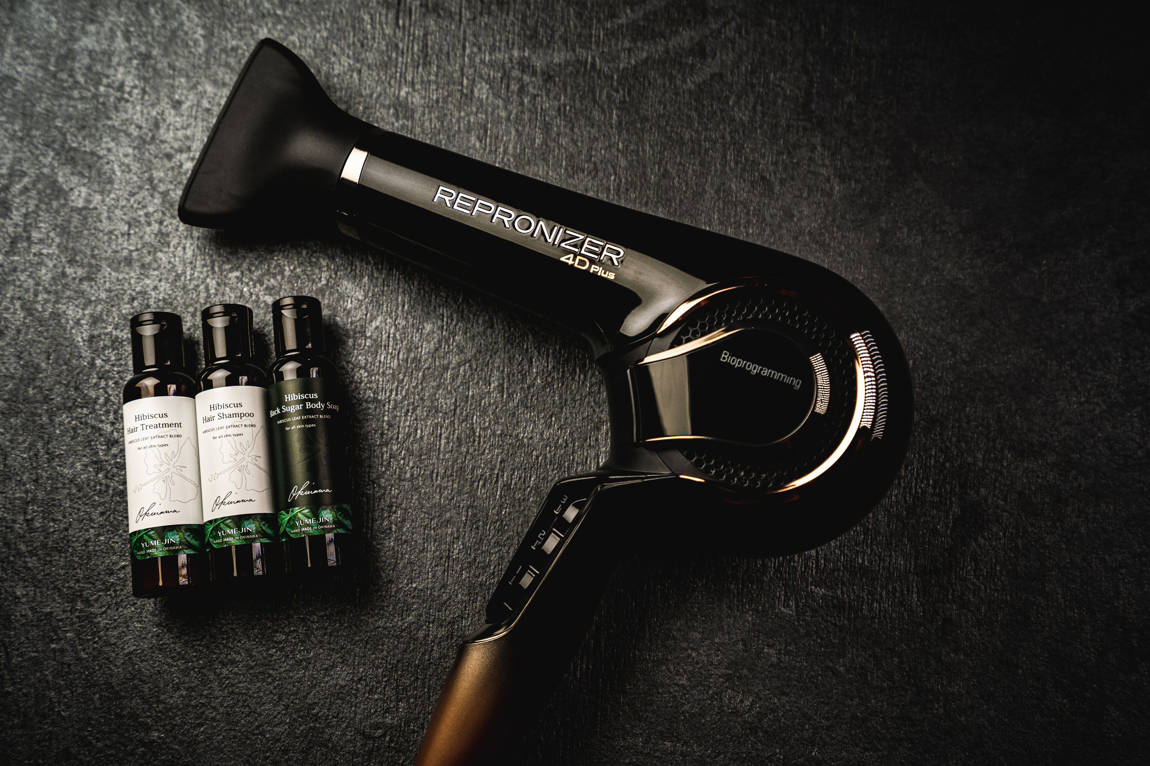 A hairdryer and three bottles of hair care products on a textured surface.