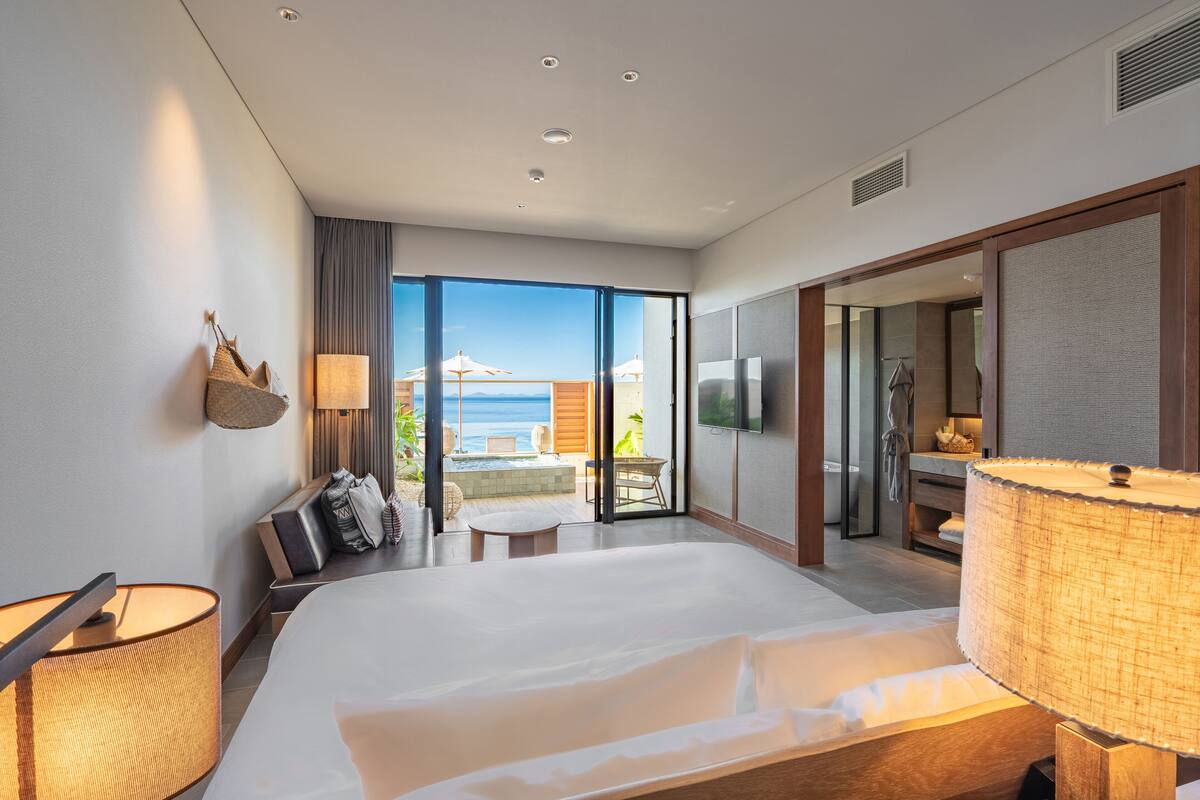A modern hotel room with a large bed, a sitting area with a view of the sea, and a balcony.