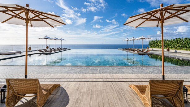 Un espace aménagé au bord de la piscine, avec des chaises longues et des parasols, offrant une vue sur un ciel bleu azur et l’océan.