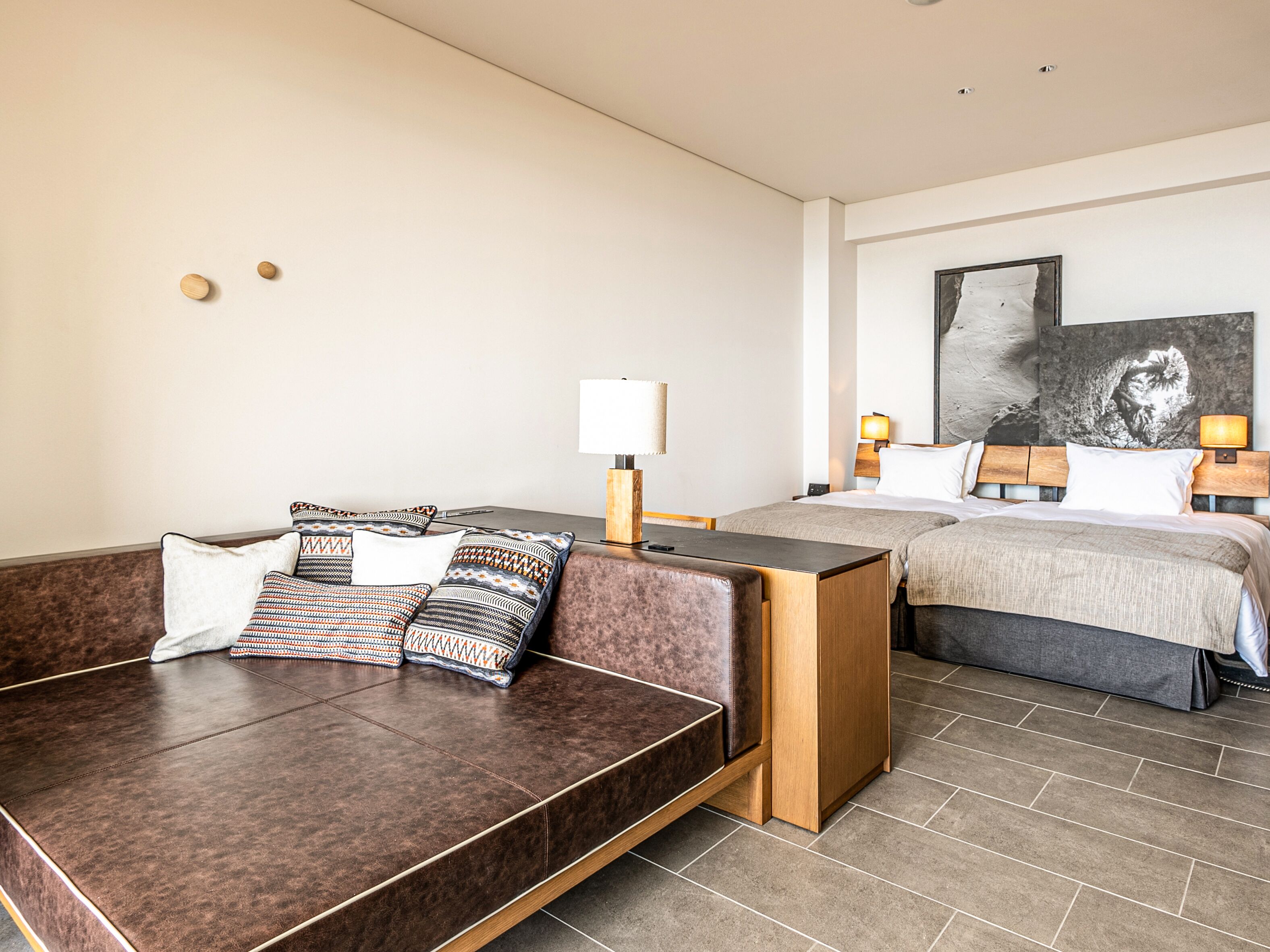 A modern hotel room with a brown leather sofa, a wooden side table, a bed with a headboard, and a large abstract painting on the wall.