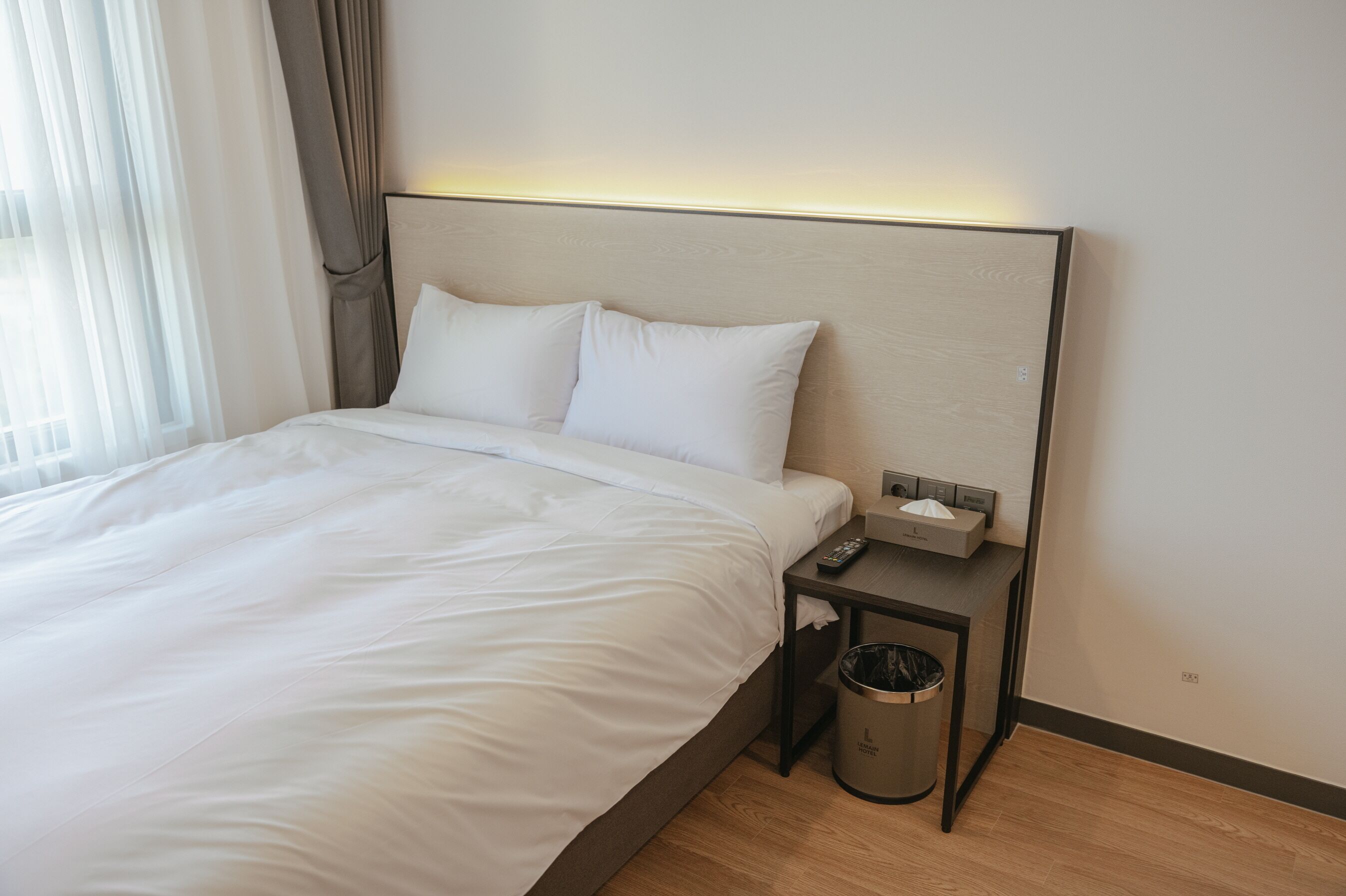 Une chambre d’hôtel moderne avec un grand lit, une table de chevet avec une télécommande, une poubelle et une lampe fixée au mur.