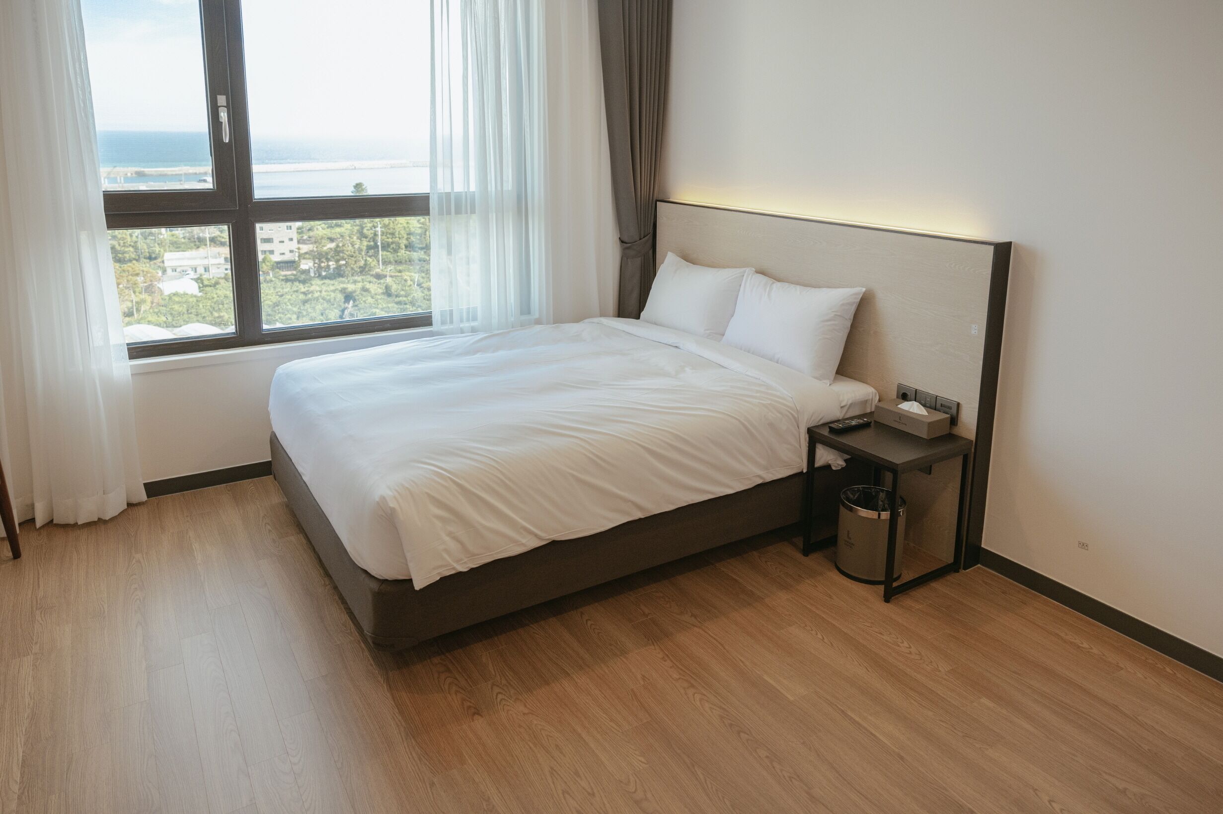 A neatly made bed in a hotel room with a view of the sea through a large window.