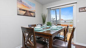 A dining area with a wooden table set for six, a painting on the wall, and a view through a large window.