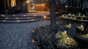 Un patio empedrado con iluminación exterior, un área de estar cubierta y un edificio con entrada visible.
