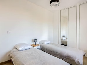 A minimalist bedroom with two single beds, a bedside table with a lamp, and a large mirror.