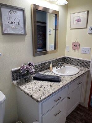 Un baño con encimera de mármol, un lavamanos, un espejo y un cartel enmarcado que dice "Amazing Grace".