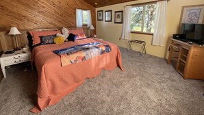 Un dormitorio acogedor con techo de madera, una cama grande con manta colorida, un mueble para televisión y una ventana con cortinas.