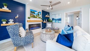A modern living room with a fireplace, a ceiling fan, and a large window with a view of palm trees.