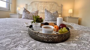 Una cama bien hecha con una bandeja de desayuno, que incluye frutas, pastelería y una taza de café, en una habitación con ropa de cama estampada y almohadones decorativos.
