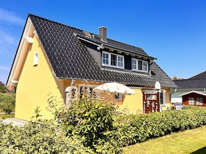 Ein gelbes Haus mit schwarzem Ziegeldach, einem sichtbaren Schornstein und einer kleinen Veranda mit einem weißen Sonnenschirm.