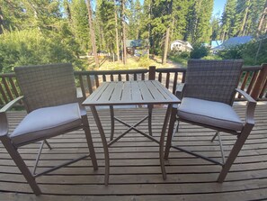 Une terrasse en bois avec deux fauteuils en osier et une table, entourée d’une zone boisée.