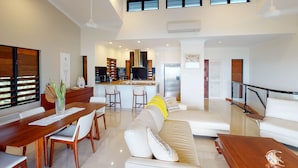 A spacious living area with a dining table, sofa, and coffee table. A kitchen with a central island and bar stools is visible in the background. - Anchorage 14, Anchorage 14 on Hamilton Island (Hamilton Island)