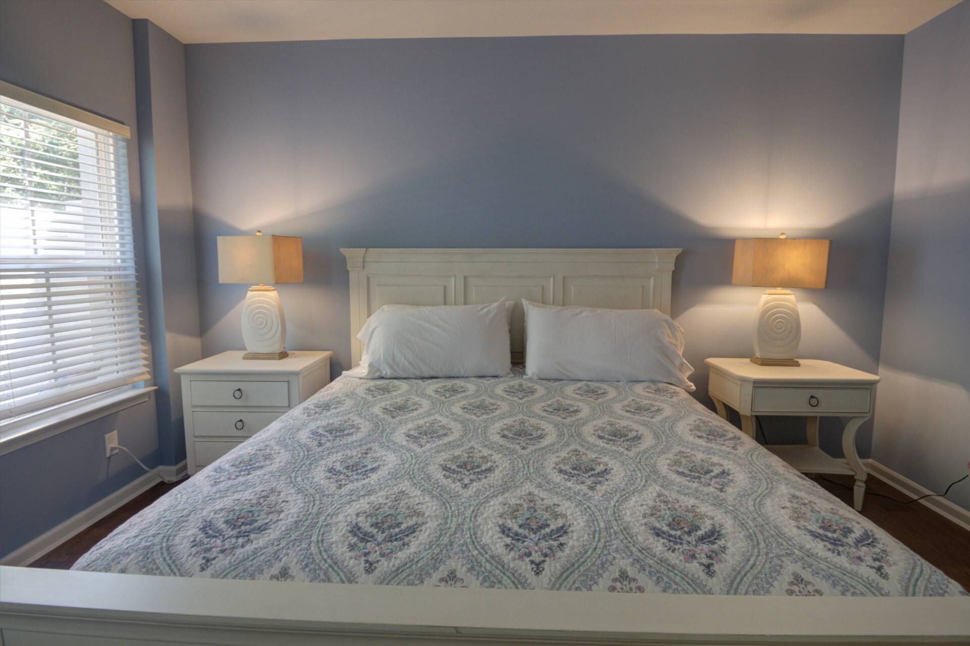 Una cama bien hecha con una colcha estampada, dos mesitas de noche con lámparas y una ventana con persianas.